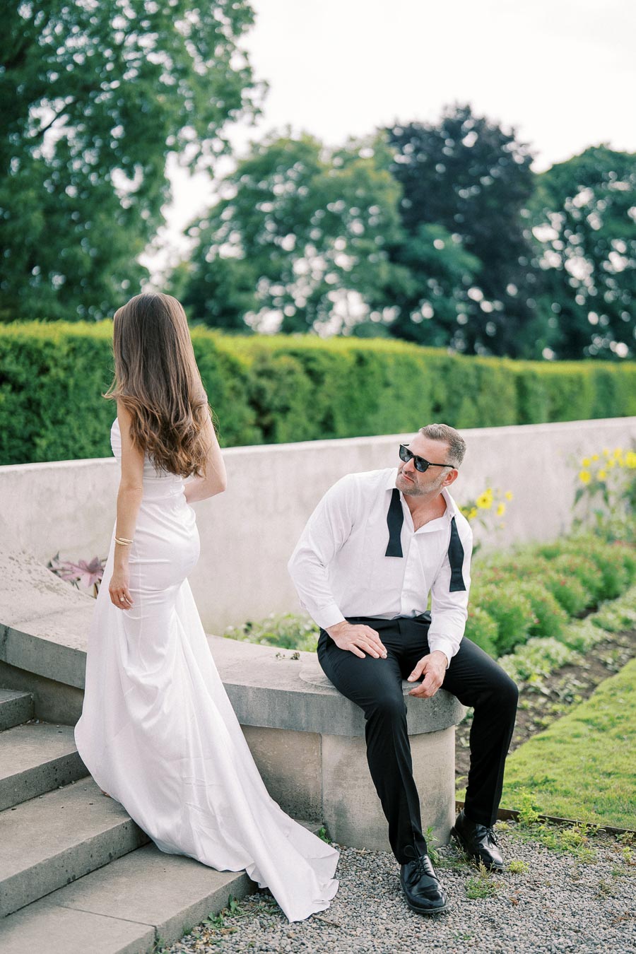 A stylish couple in formal attire sits on stone steps in a lush garden, with the woman in a white dress and the man wearing sunglasses and a loosened bow tie.