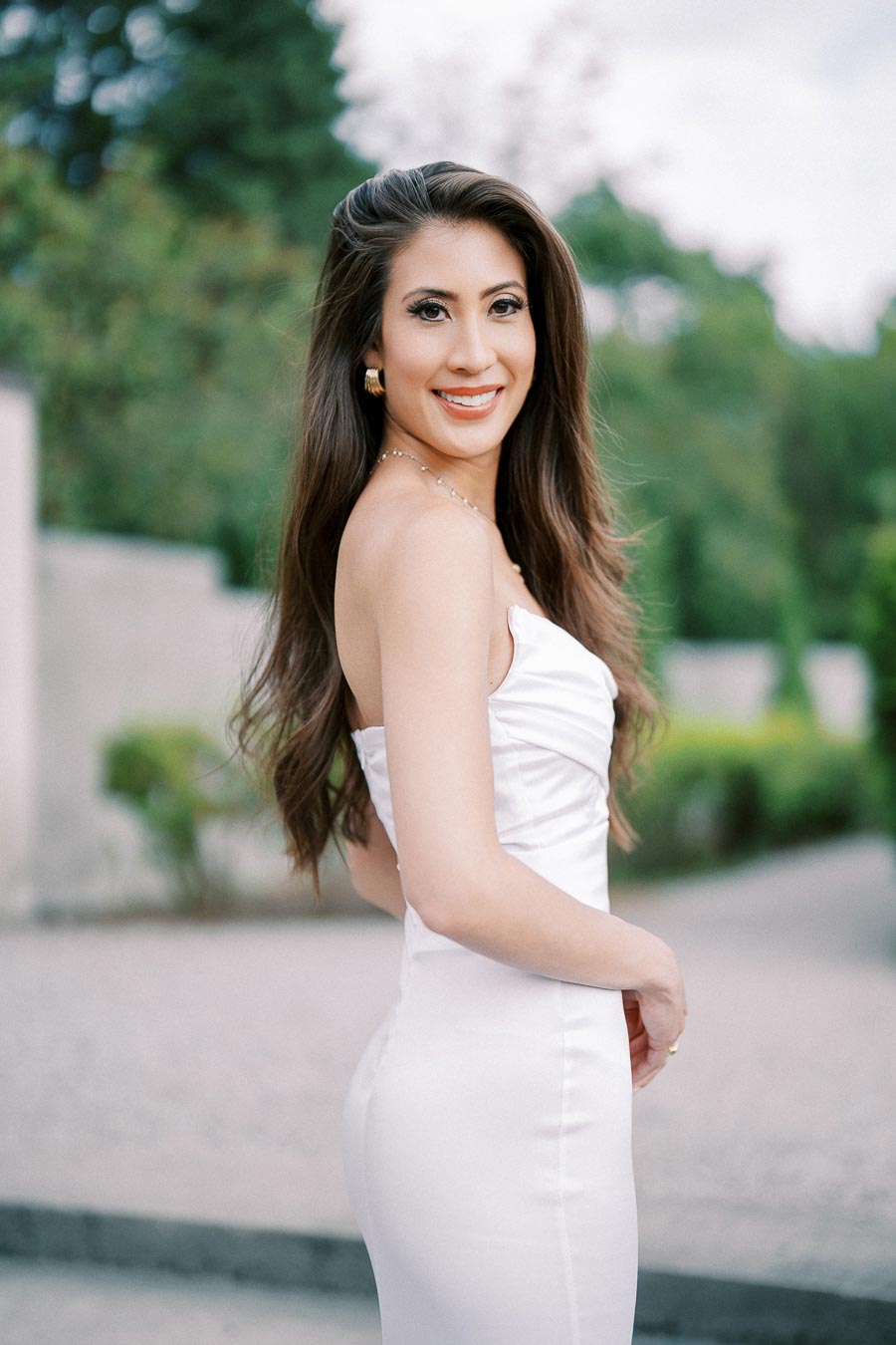 A woman in a white dress smiling outdoors with a blurred green background.