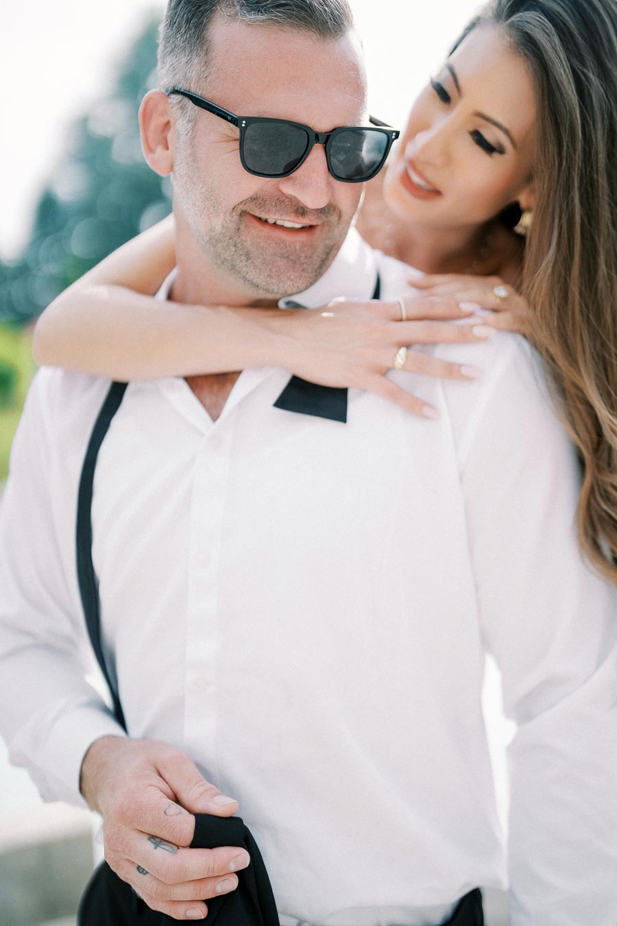 A smiling couple enjoying a sunny day, with a woman playfully embracing a man from behind; he is wearing sunglasses and a white shirt, showcasing a relaxed and joyful moment.