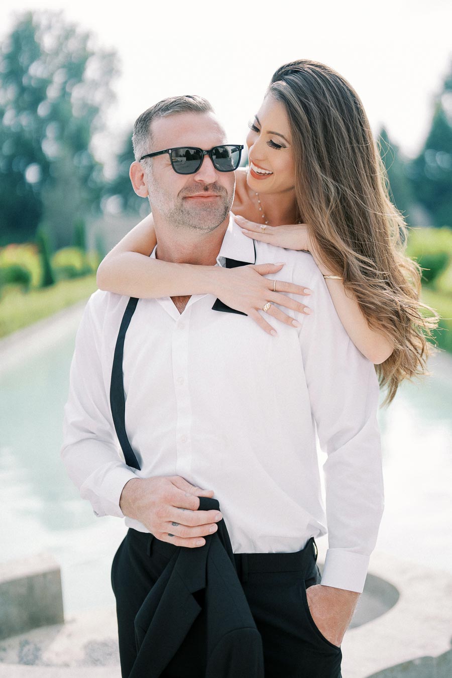 Happy couple posing outdoors, woman playfully embracing man from behind, both dressed in white and black attire, enjoying a sunny day by the pool, capturing a moment of joy and affection.