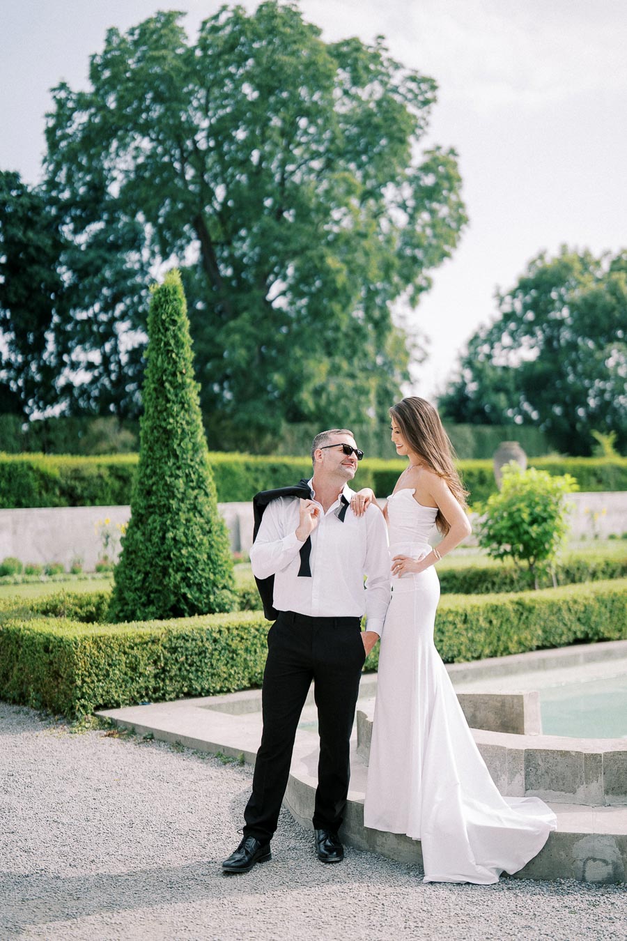 Man in a formal white shirt and black pants stands beside a woman in a white gown in a well-manicured garden setting, with lush green trees and hedges in the background.
