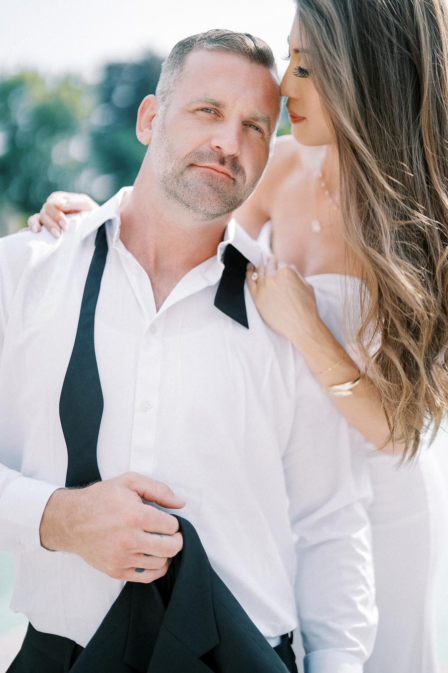 A couple embracing outdoors with the man in a white shirt and untied black bow tie, and the woman in a strapless dress, both looking content and affectionate.