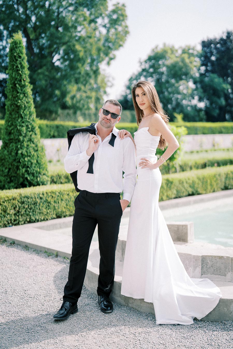 A stylish couple poses outdoors on a bright day. The man is wearing sunglasses, a white shirt, and black pants, holding his jacket over his shoulder. The woman is in an elegant white dress, standing beside a fountain, surrounded by lush greenery.