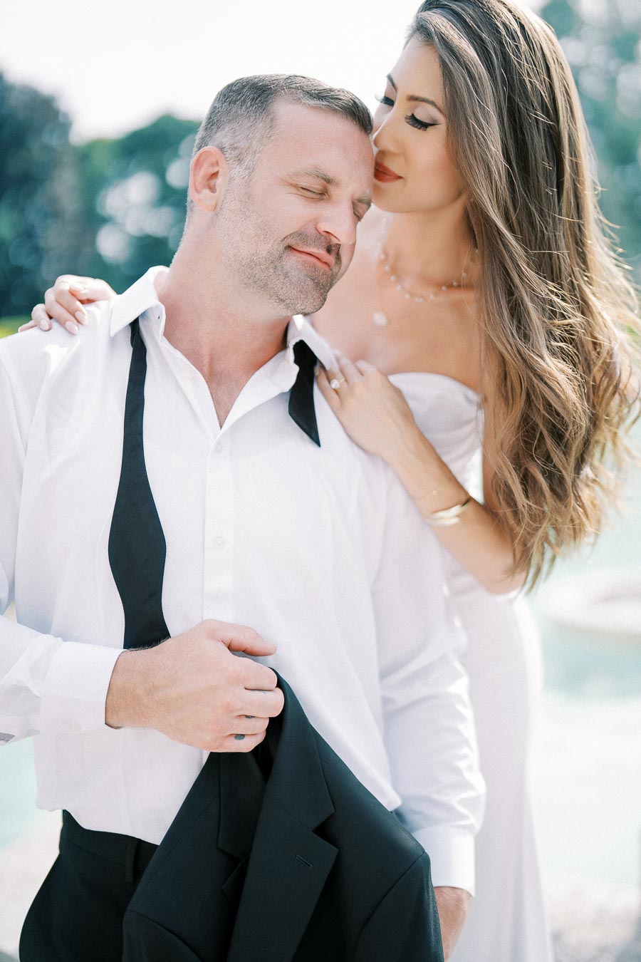 A couple embraces outdoors, with a man in a white shirt and untied black bow tie smiling as he holds a suit jacket, and a woman in a strapless white dress affectionately leaning on his shoulder, surrounded by a blurred natural background.