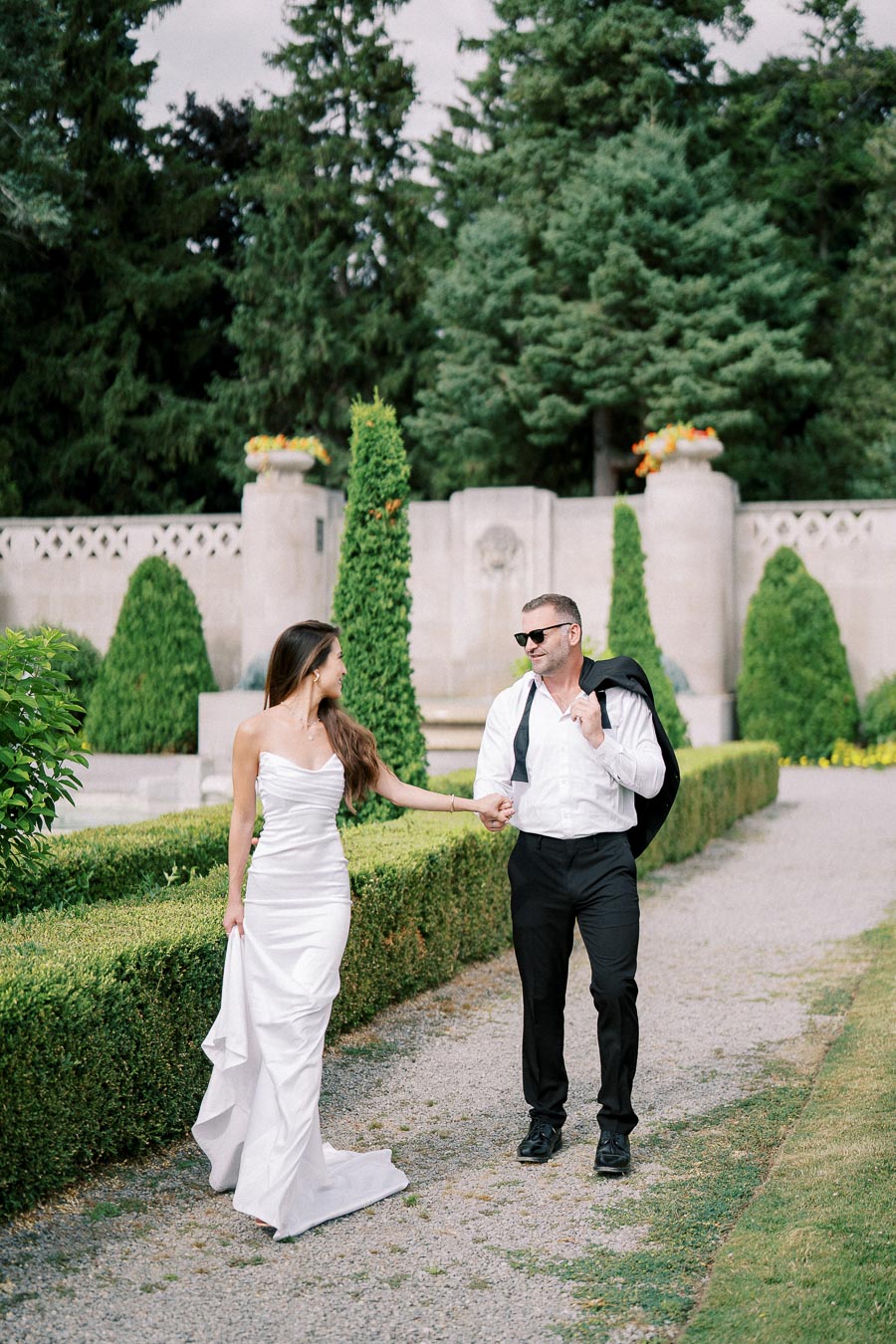 A couple walks hand-in-hand through a beautifully manicured garden, with tall trees and elegant architecture in the background; the woman is wearing a white dress, and the man is dressed in a white shirt and dark pants.