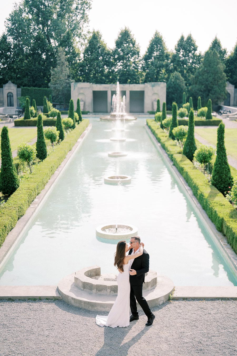 A romantic couple embraces beside a long, elegant reflecting pool with fountains, surrounded by manicured gardens and tall hedges, under a clear sky.