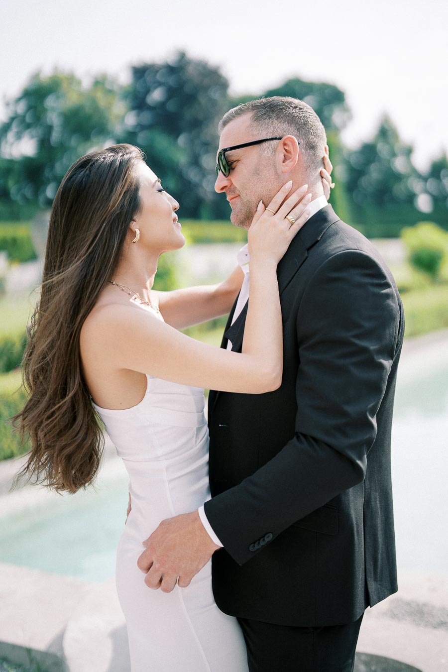A couple in elegant attire embracing outdoors by a serene pool, surrounded by lush greenery, exuding romance and joy.
