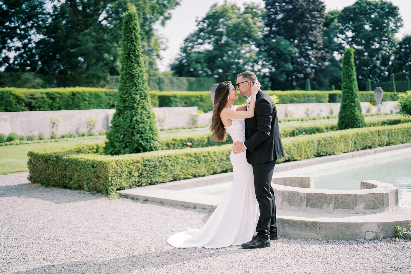 A bride and groom embrace lovingly in a beautifully manicured garden, standing next to a decorative pool, surrounded by lush greenery and topiary.