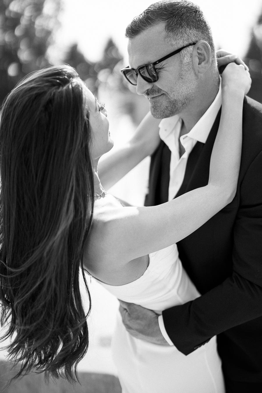 A black and white photo of a couple embracing, with a woman in a white dress and a man in a suit wearing sunglasses, sharing an intimate moment outdoors.