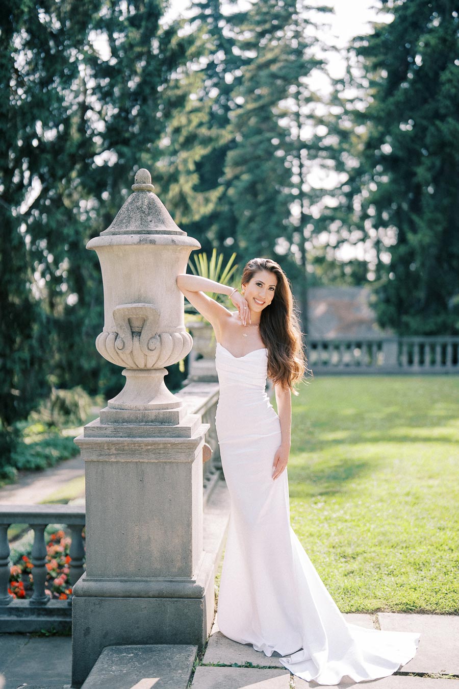 A bride in an elegant white wedding gown poses next to a stone urn in a lush garden, surrounded by vibrant greenery and sunlight.