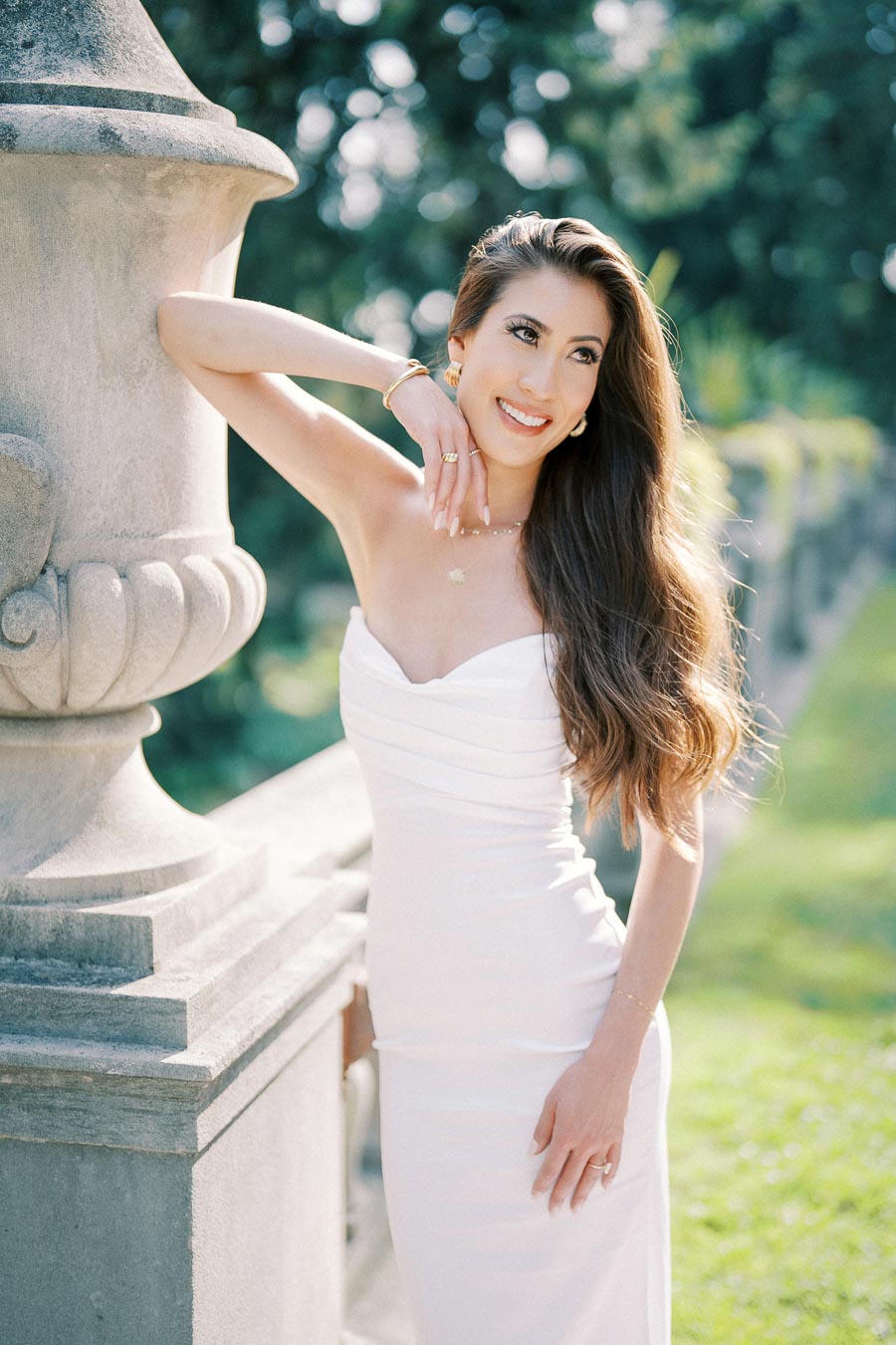 A woman in an elegant white strapless dress smiles while leaning against a stone structure in a sunlit garden setting.