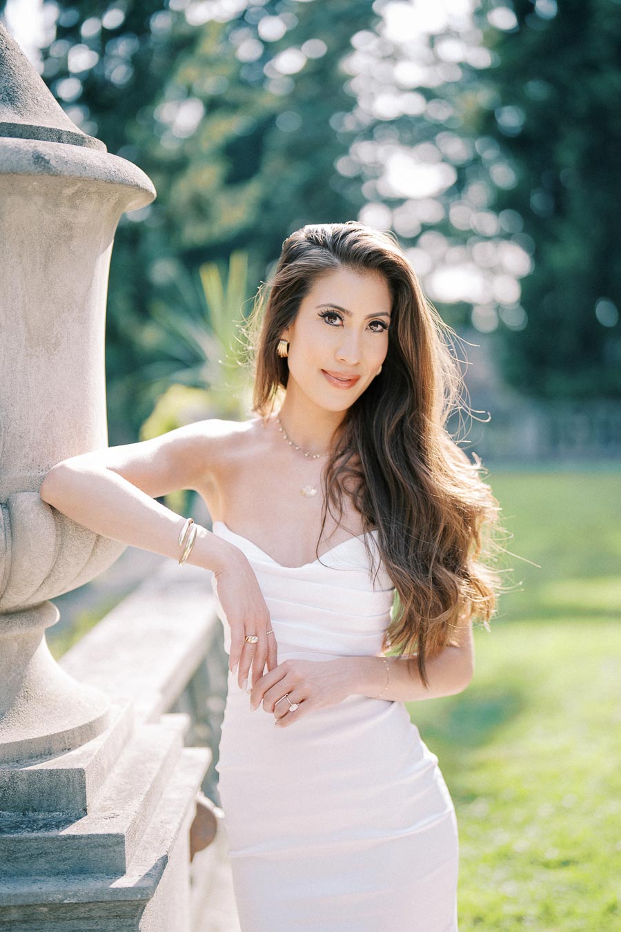 Elegant woman in a white dress leaning against a stone railing outdoors, with a serene garden backdrop and soft sunlight highlighting her long hair