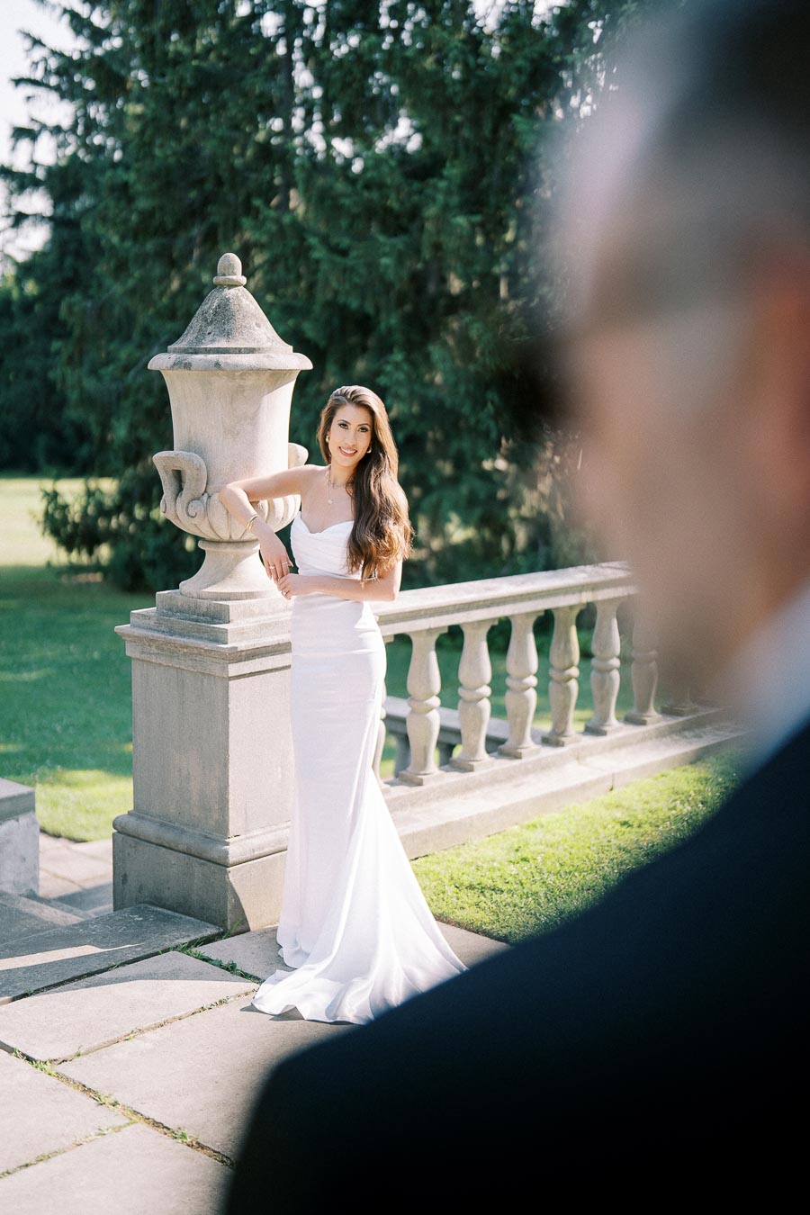 Bride in elegant white gown posing by a stone balustrade outdoors with lush green trees in the background on a sunny day.