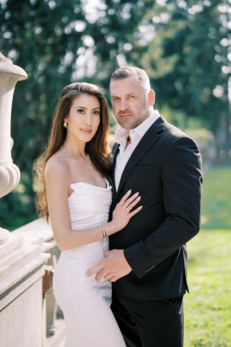 A couple posing elegantly in formal attire, with the woman in a white dress and the man in a black suit, set against a scenic outdoor backdrop with greenery.