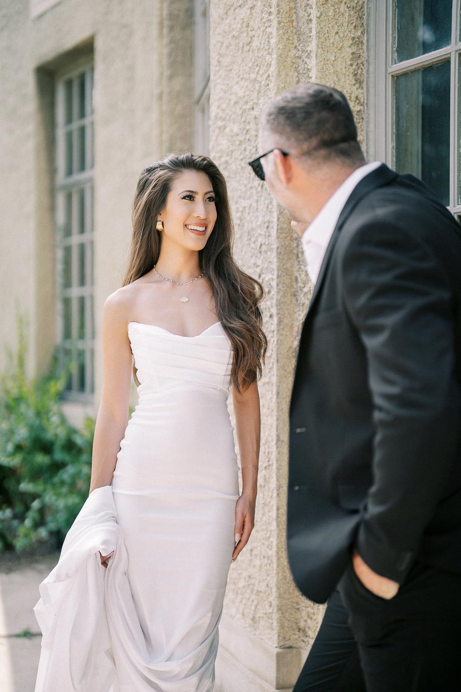 Elegant woman in a strapless white dress smiling at a man in a suit, standing near a rustic building with greenery in the background.