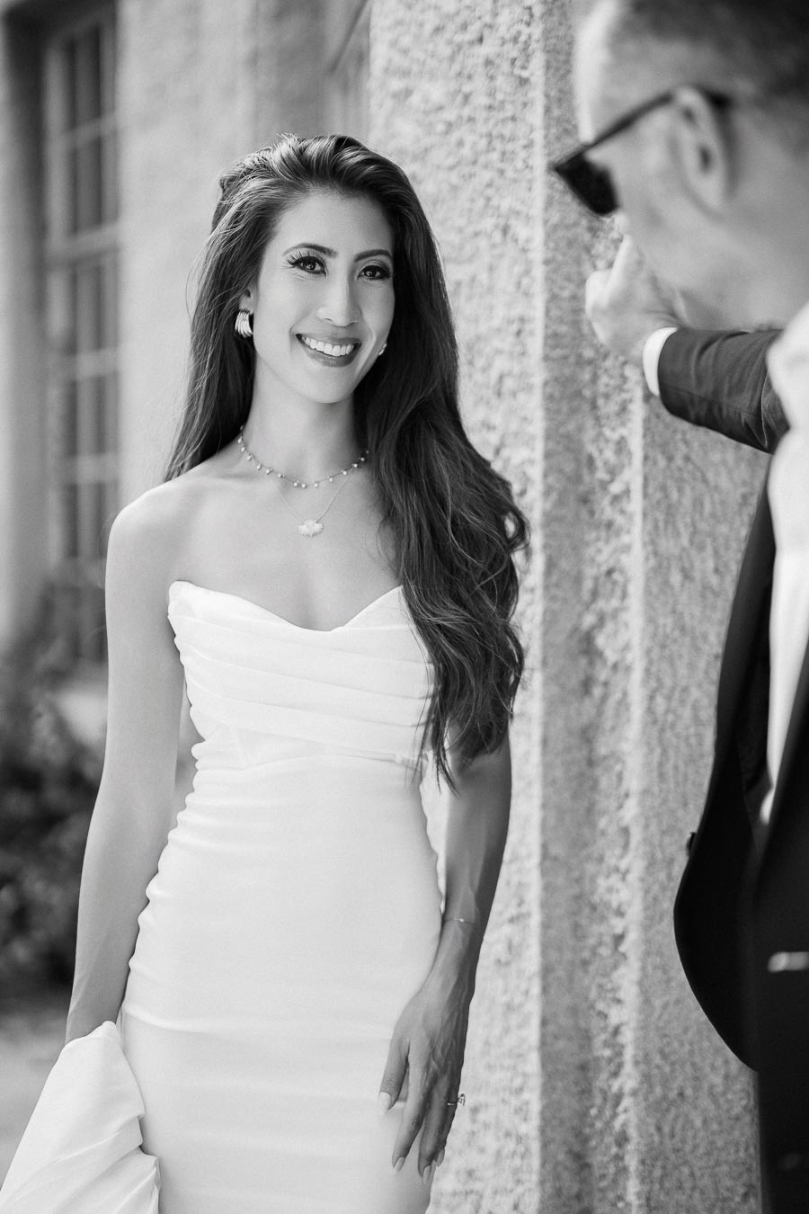 A bride in a strapless white wedding gown smiling warmly, standing against a textured wall, with a man in a suit partially visible. Black and white photograph capturing a joyful wedding moment.