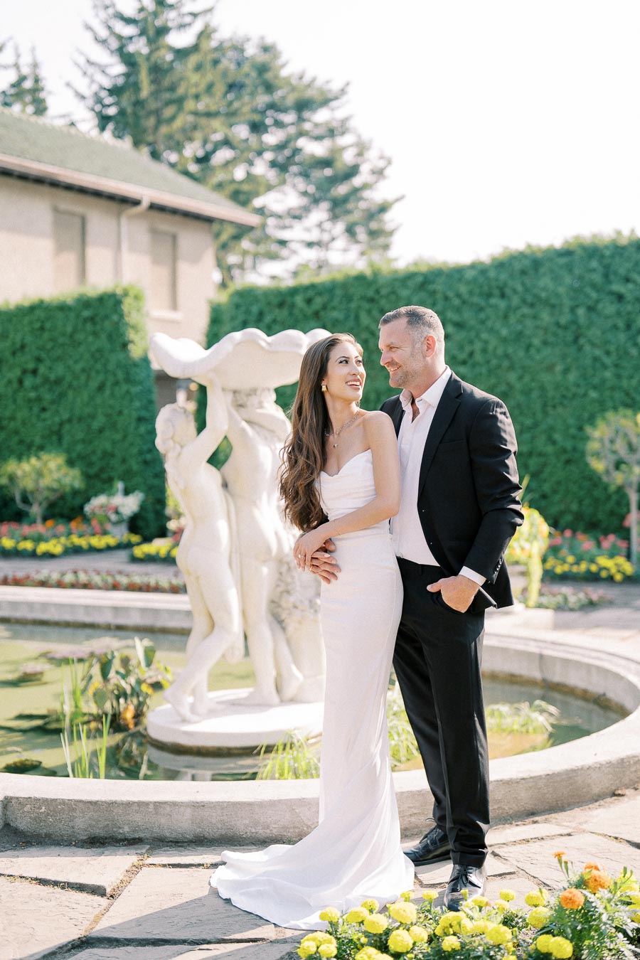 A couple smiling and embracing in elegant wedding attire near a classic stone fountain, surrounded by vibrant garden scenery.