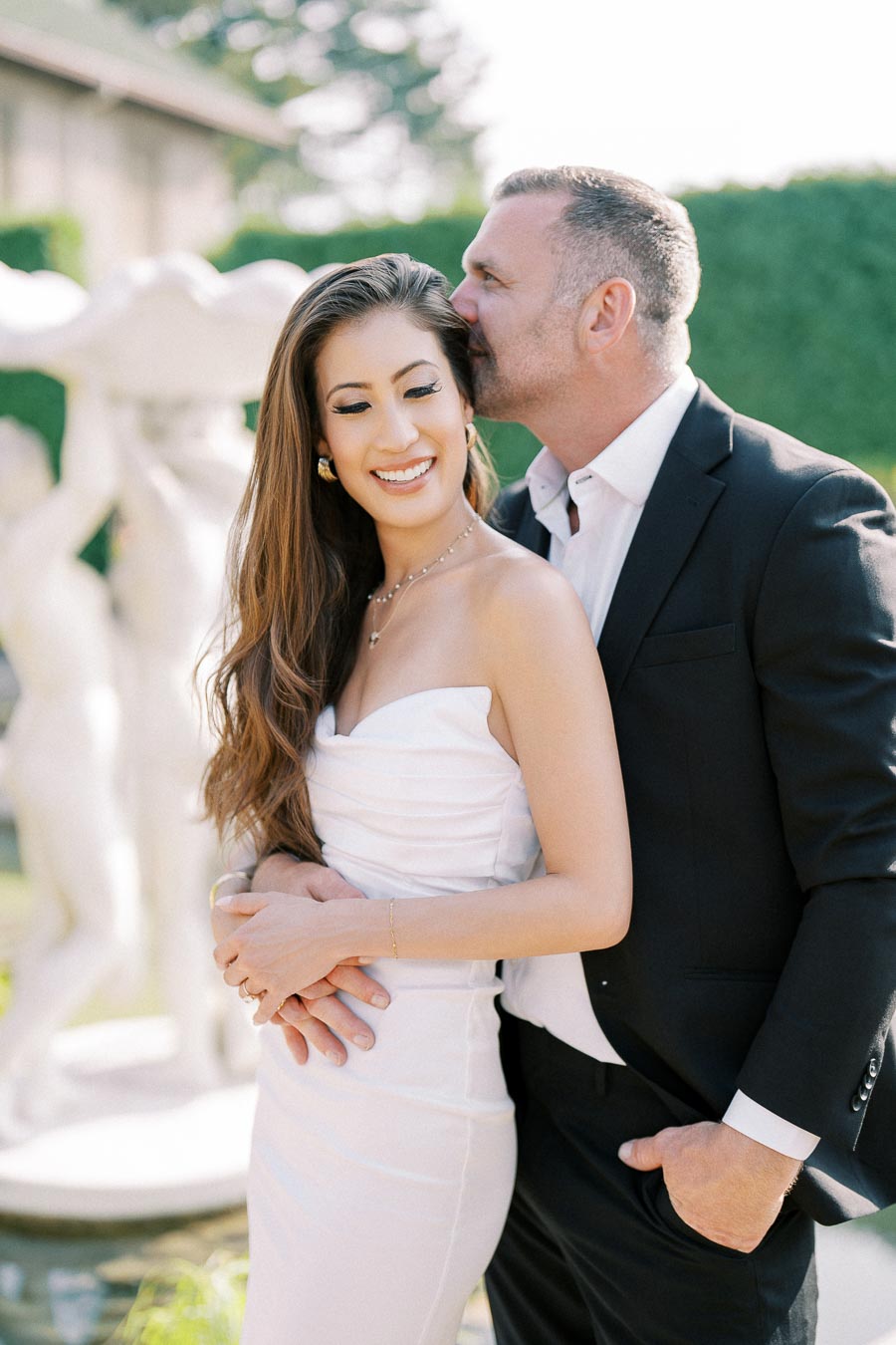 A couple dressed elegantly in a garden setting, with the man lovingly embracing the woman from behind, both smiling.
