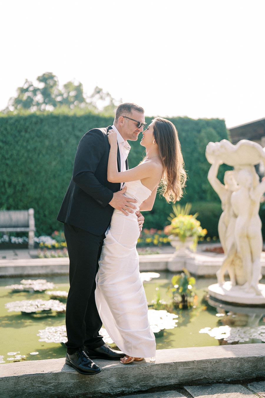 A couple in elegant attire embraces romantically by a serene garden pond with statues and lush greenery in the background, capturing a moment of love and joy.