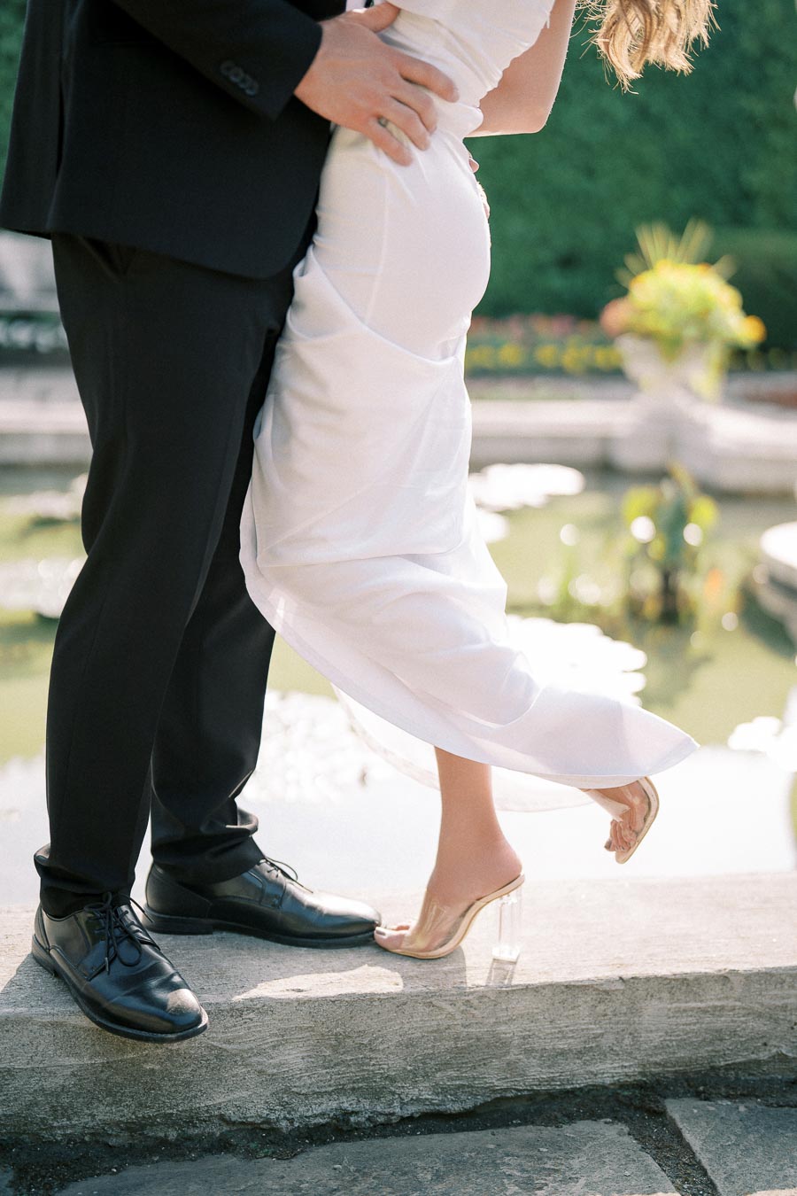 A romantic couple in formal attire embracing by a serene water feature, with the woman lifted slightly, her wedding dress flowing elegantly and her feet in transparent high heels.