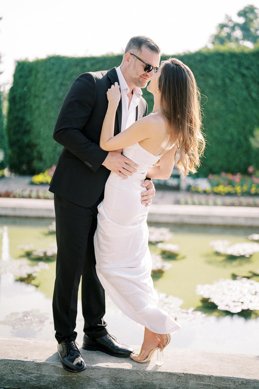 A couple dressed in elegant attire embraces by a garden pond, showcasing romance and style in an outdoor setting.