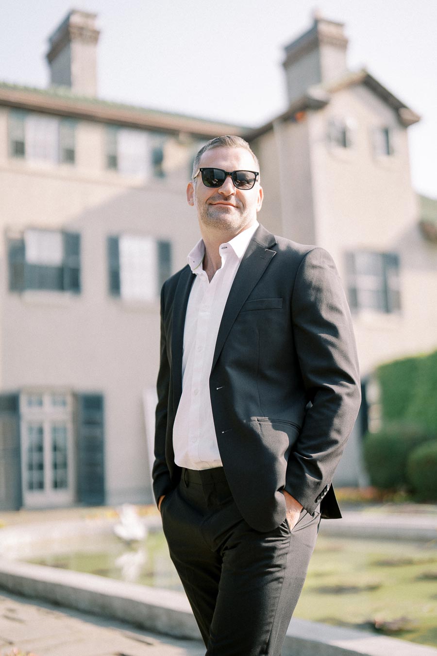 Man in a suit and sunglasses standing confidently outside a large house with a fountain in the background.