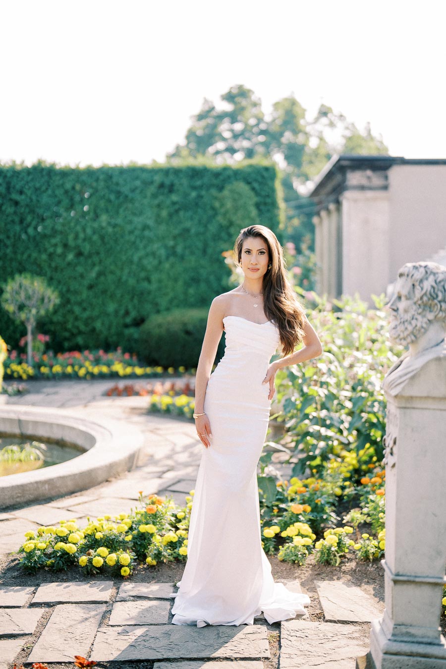 Elegant woman in a white strapless gown posing in a sunlit garden, surrounded by vibrant flowers and lush greenery, ideal for a wedding or romantic outdoor event.