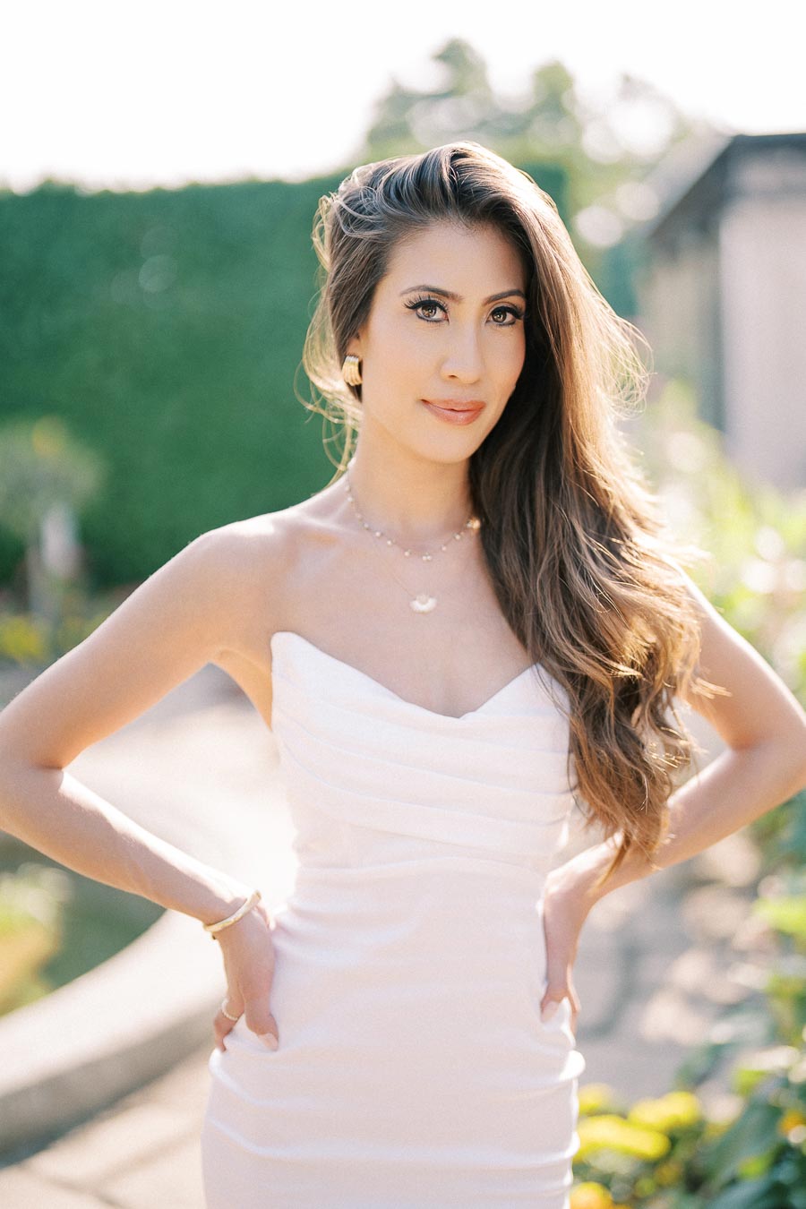 Elegant woman in a white sleeveless dress posing outdoors with long flowing hair, surrounded by greenery and sunlight