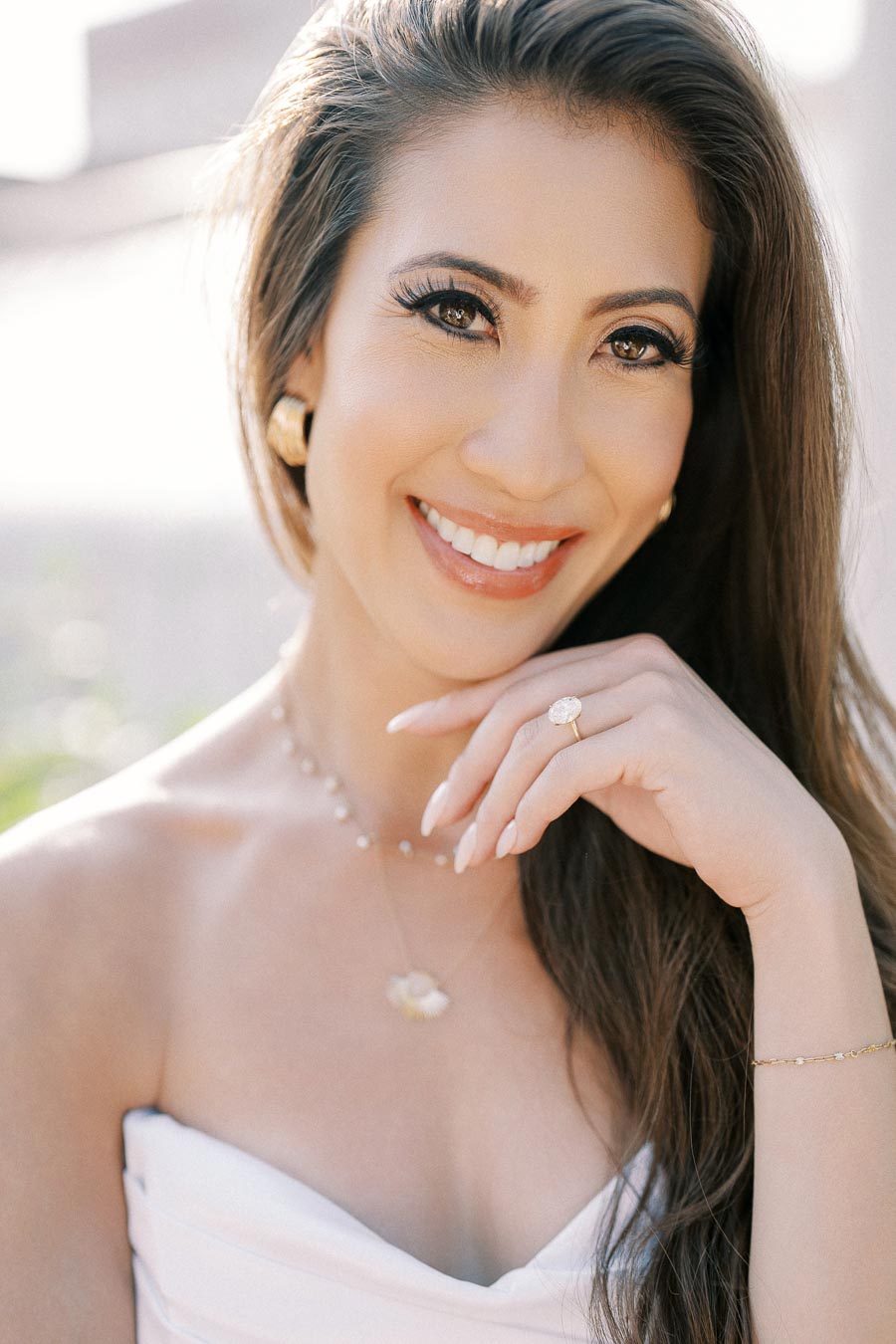 Smiling woman with long hair wearing a strapless white top, accessorized with gold earrings, necklace, and a ring, posing outdoors in bright, natural lighting.
