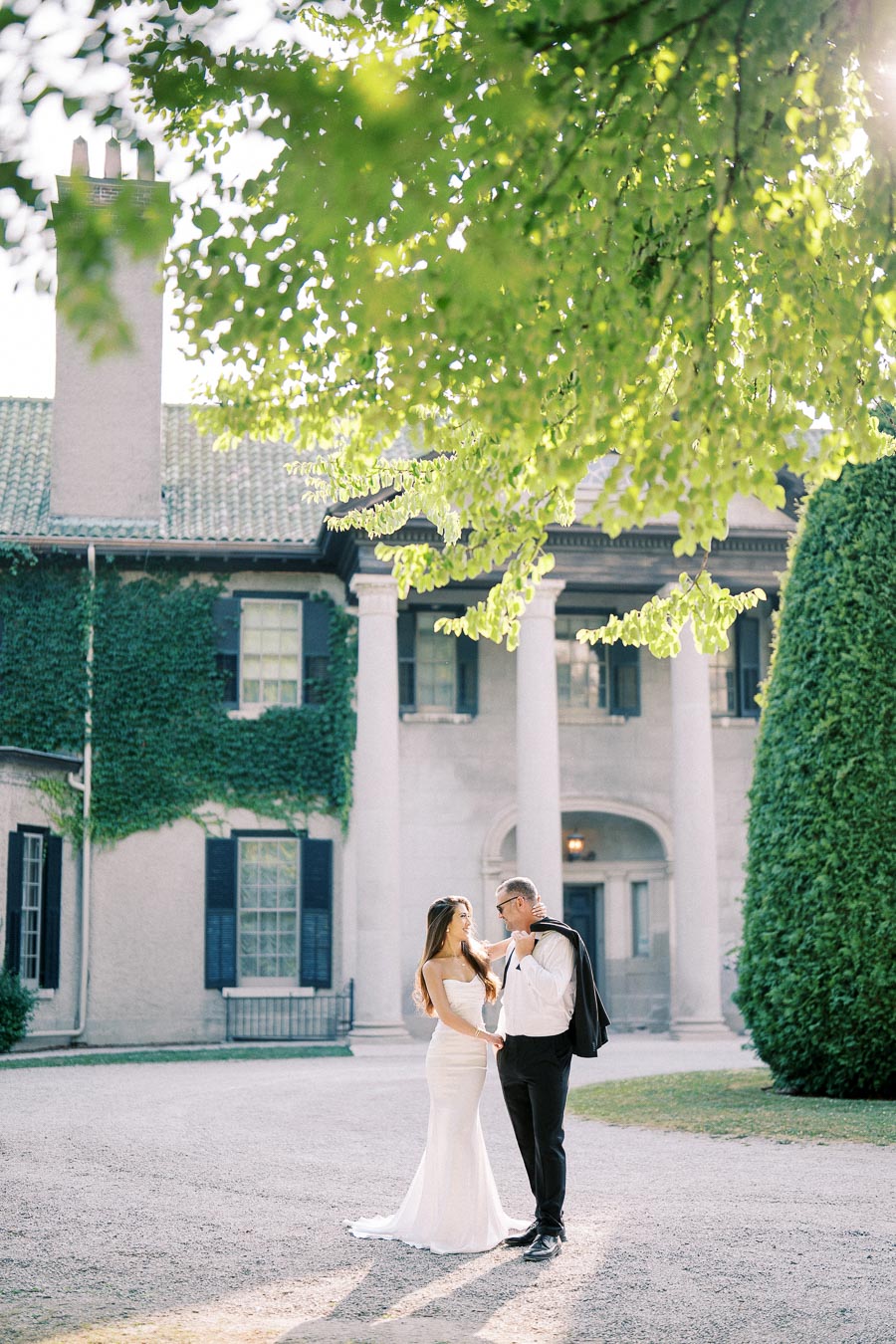 A bride and groom standing affectionately in front of an elegant mansion covered in ivy, surrounded by lush greenery, on a sunny day.