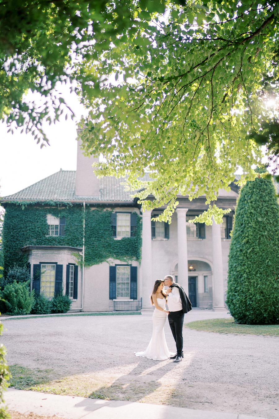 A romantic couple kissing in front of an ivy-covered mansion under lush green tree branches.