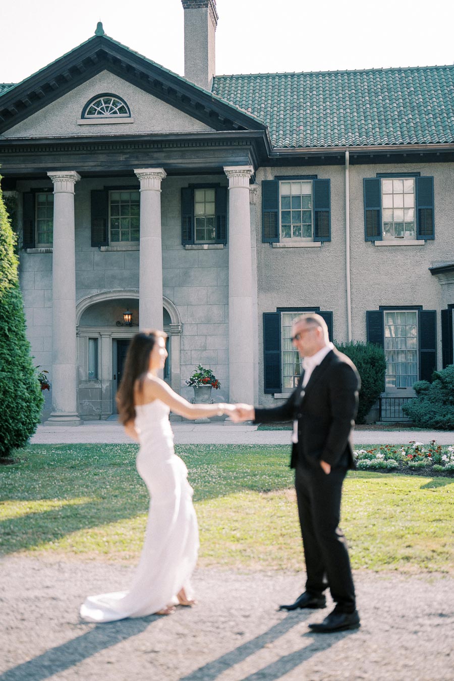 A couple elegantly dressed in formal attire shares a dance in front of a grand, historic mansion with large columns and a tiled roof, surrounded by lush greenery and floral arrangements on a sunny day.