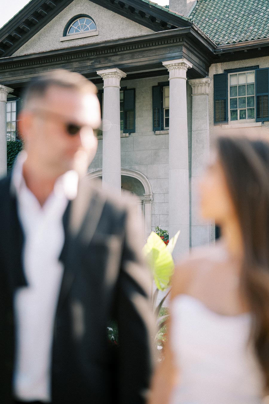 Blurred image of a couple standing in front of an elegant historic mansion with tall white columns and a gabled roof, highlighting classic architectural features.