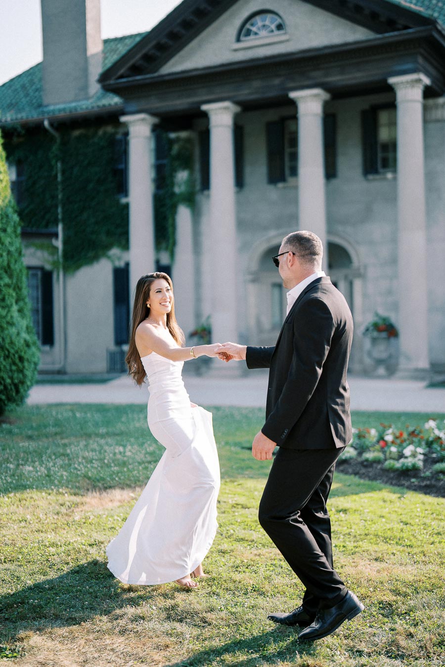 A couple elegantly dancing in front of a historic mansion with columns, with the woman in a white dress and the man in a black suit, surrounded by a lush garden and clear sky.