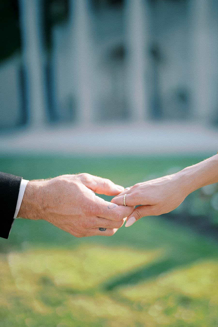 A close-up of a couple holding hands during a wedding ceremony, with one person wearing a gold wedding ring, set against a green outdoor background.