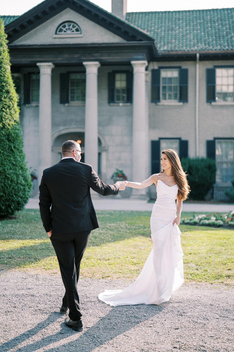 Elegant bride in a white wedding gown and groom in a black suit dancing in front of a grand mansion with columns, creating a romantic and timeless wedding scene.