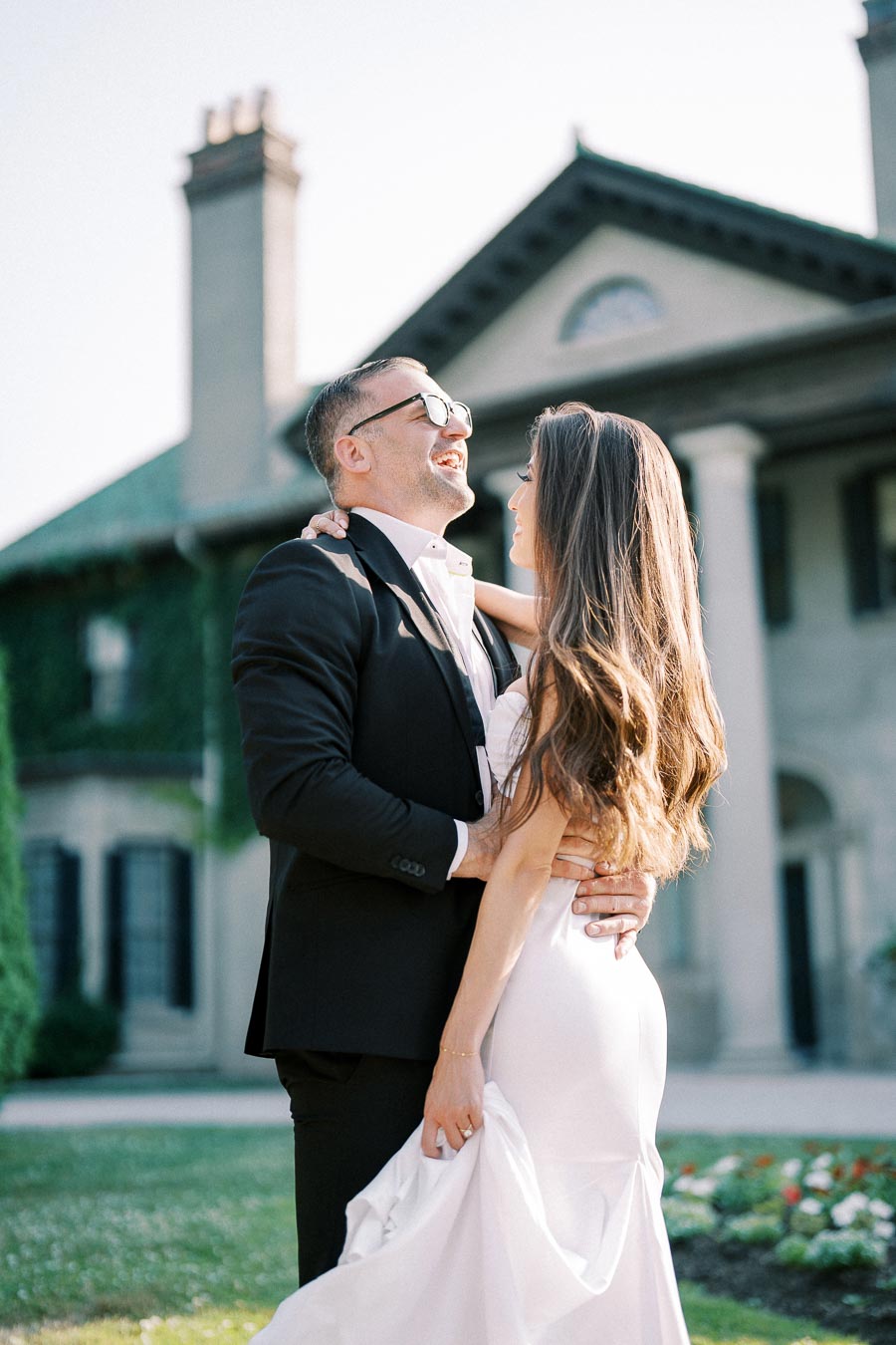 A couple in formal attire shares a joyful moment outside a historic mansion. The man is wearing a black suit and sunglasses, while the woman is in a flowing white dress, set against a background of greenery and elegant architecture.
