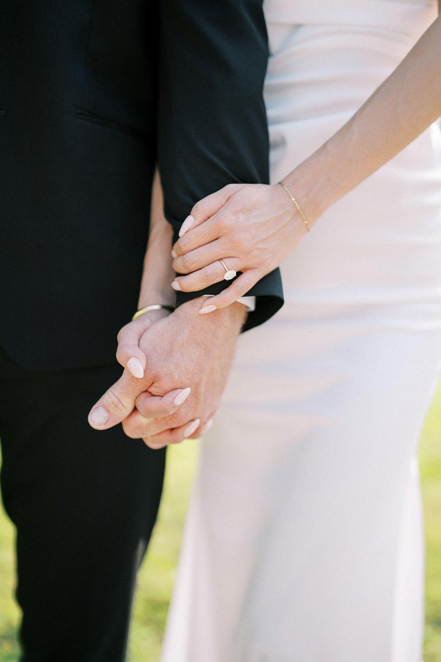Close-up of a couple holding hands, showcasing an engagement ring on the woman's hand, with the man dressed in a black suit and the woman in a white dress, symbolizing love and commitment.