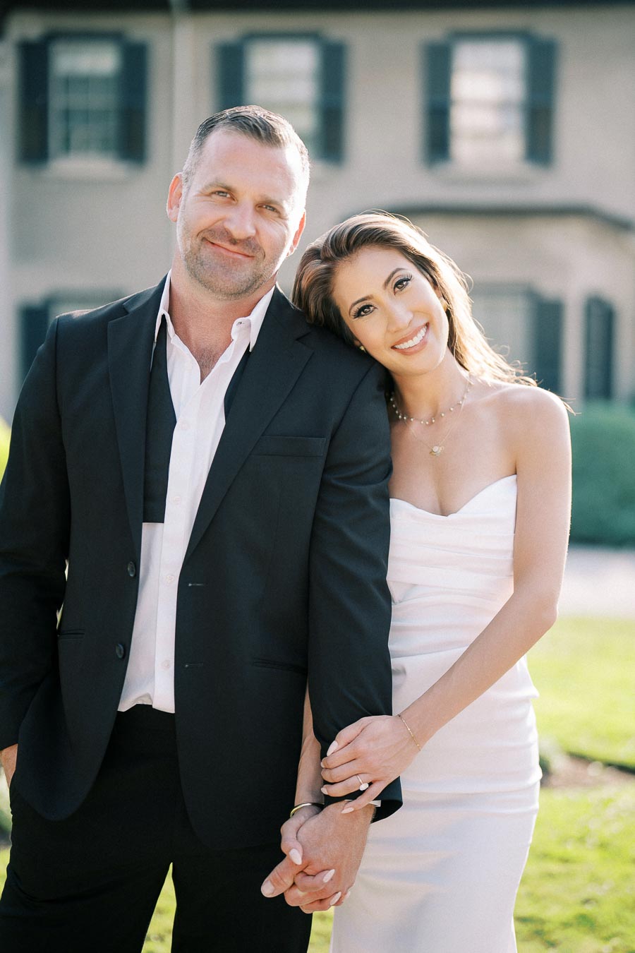 A happy couple standing closely together in front of a house, with the woman smiling and resting her head on the man's shoulder, both dressed in formal attire.