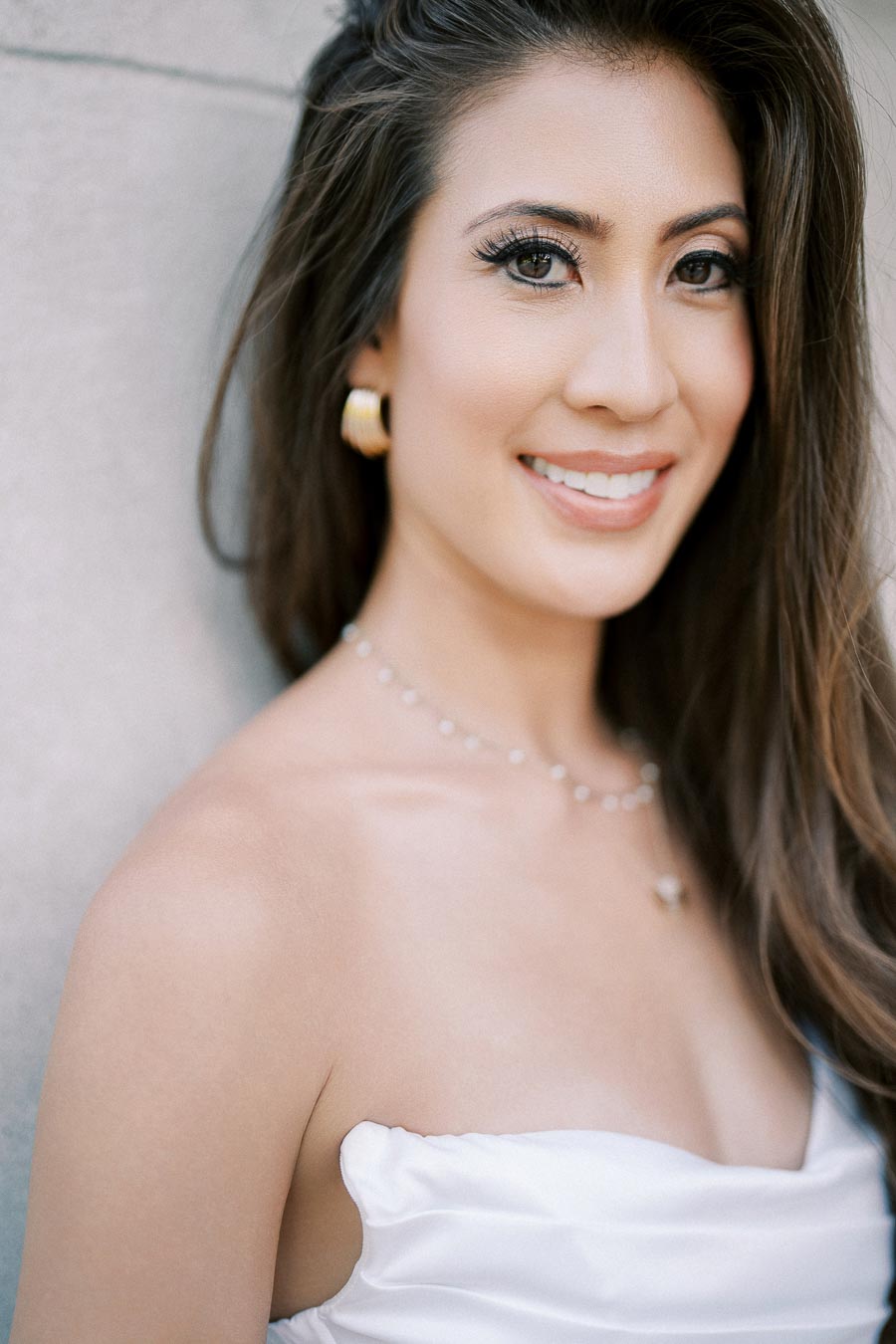 A woman with long brown hair smiling while wearing a strapless white dress and gold earrings, standing against a light-colored wall.