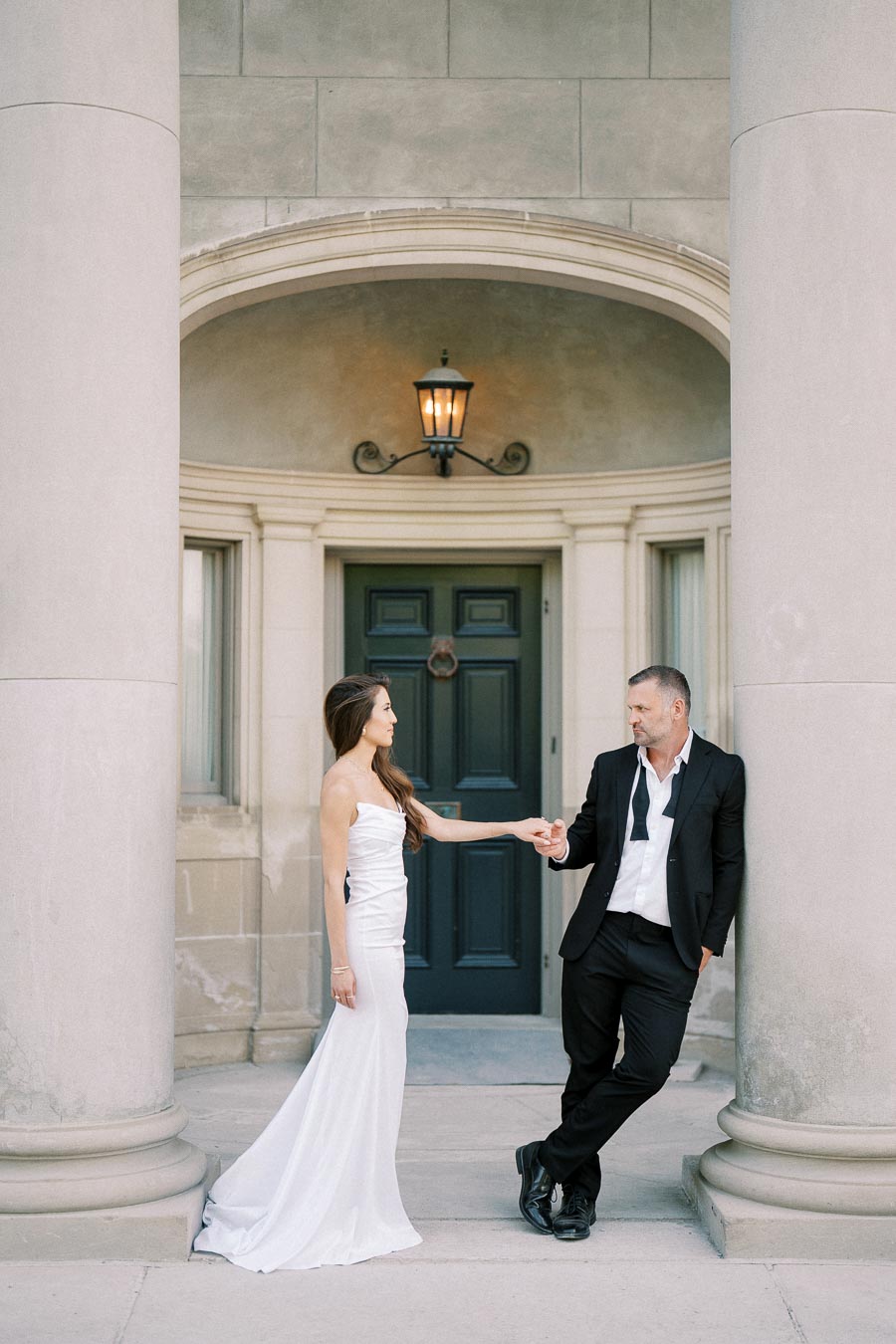 Elegant couple in formal attire standing in front of a classic building with large columns, the woman wearing a white dress and the man in a black suit with an unfastened tie, holding hands and gazing at each other.