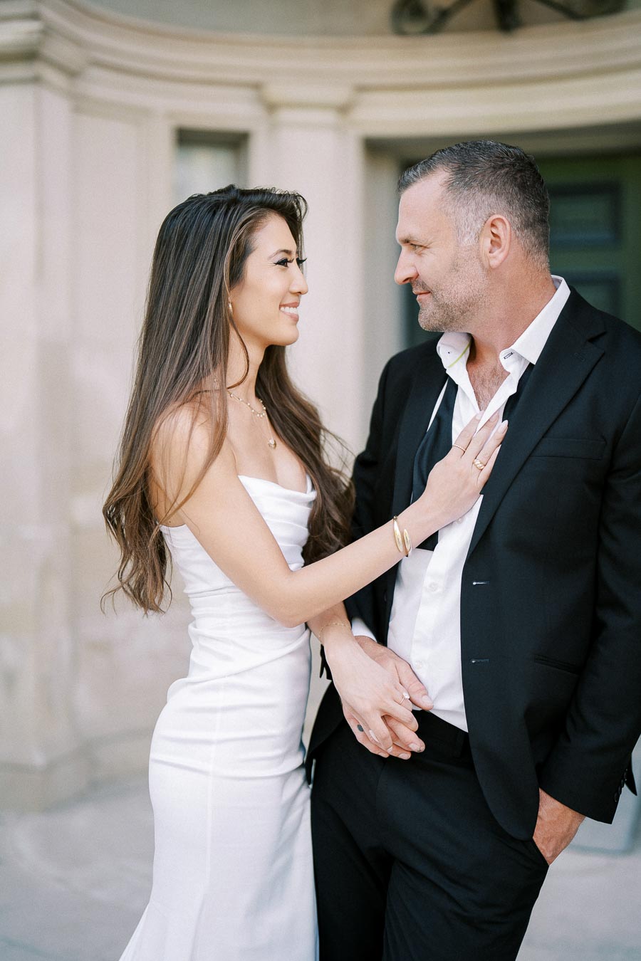 A couple in elegant attire smiling at each other, woman in white dress and man in black suit, standing outside a building.