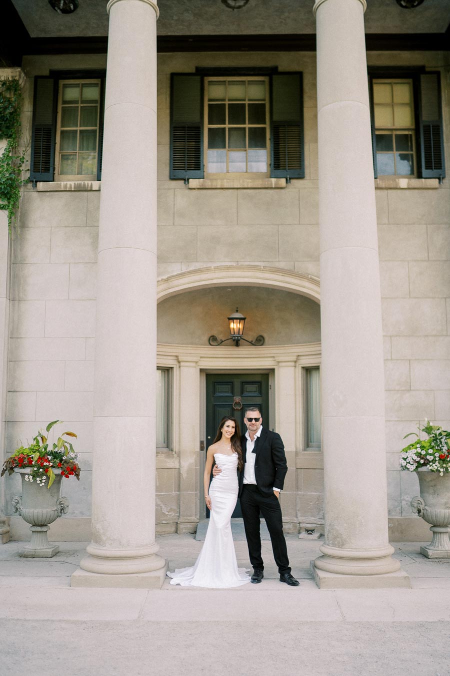 Elegant couple posing in formal attire in front of a grand stone building with tall columns and large windows.