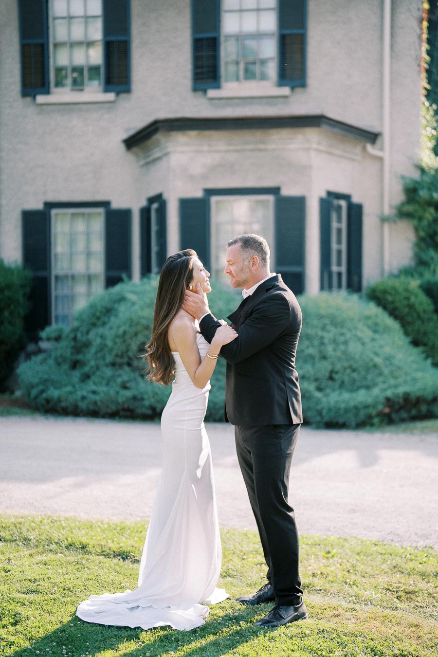 A couple dressed in elegant attire shares a romantic moment in front of a charming, historic house with green shutters and lush garden bushes, capturing a timeless wedding scene.