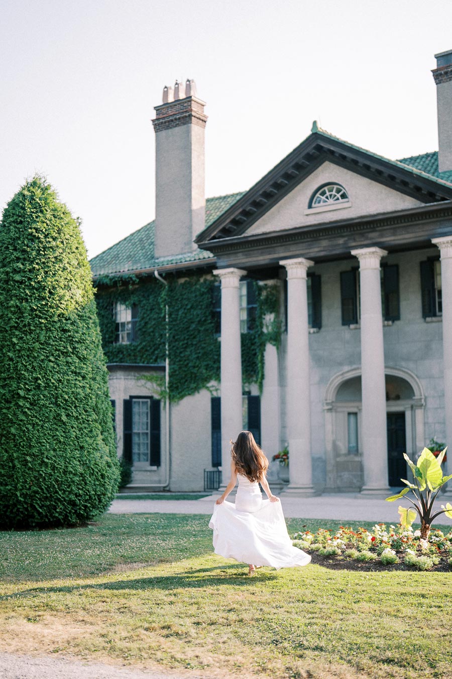Elegant woman in white dress walking towards a historic mansion covered in ivy, surrounded by lush greenery and colorful flower beds on a sunny day.