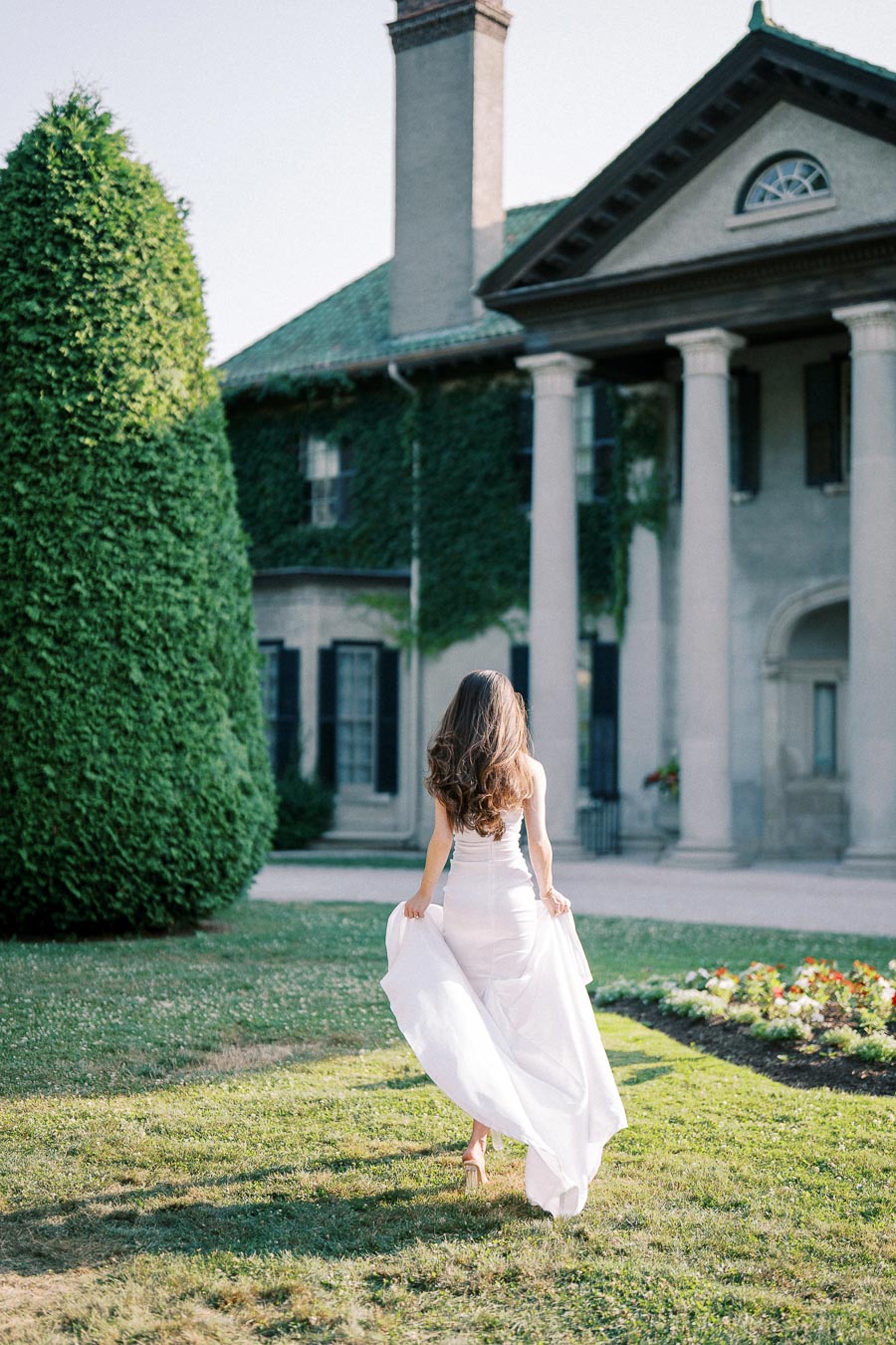 A woman in a flowing white dress walks towards a grand ivy-covered mansion with columns, surrounded by neatly manicured gardens and greenery, under a clear blue sky.