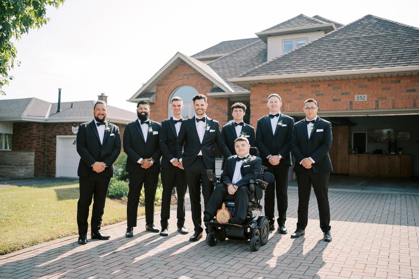 A group of seven groomsmen wearing matching black tuxedos, smiling and posing together on a sunny day in front of a brick house. One man is seated in a wheelchair, and the group is standing on a paved driveway, showcasing a classic wedding look.