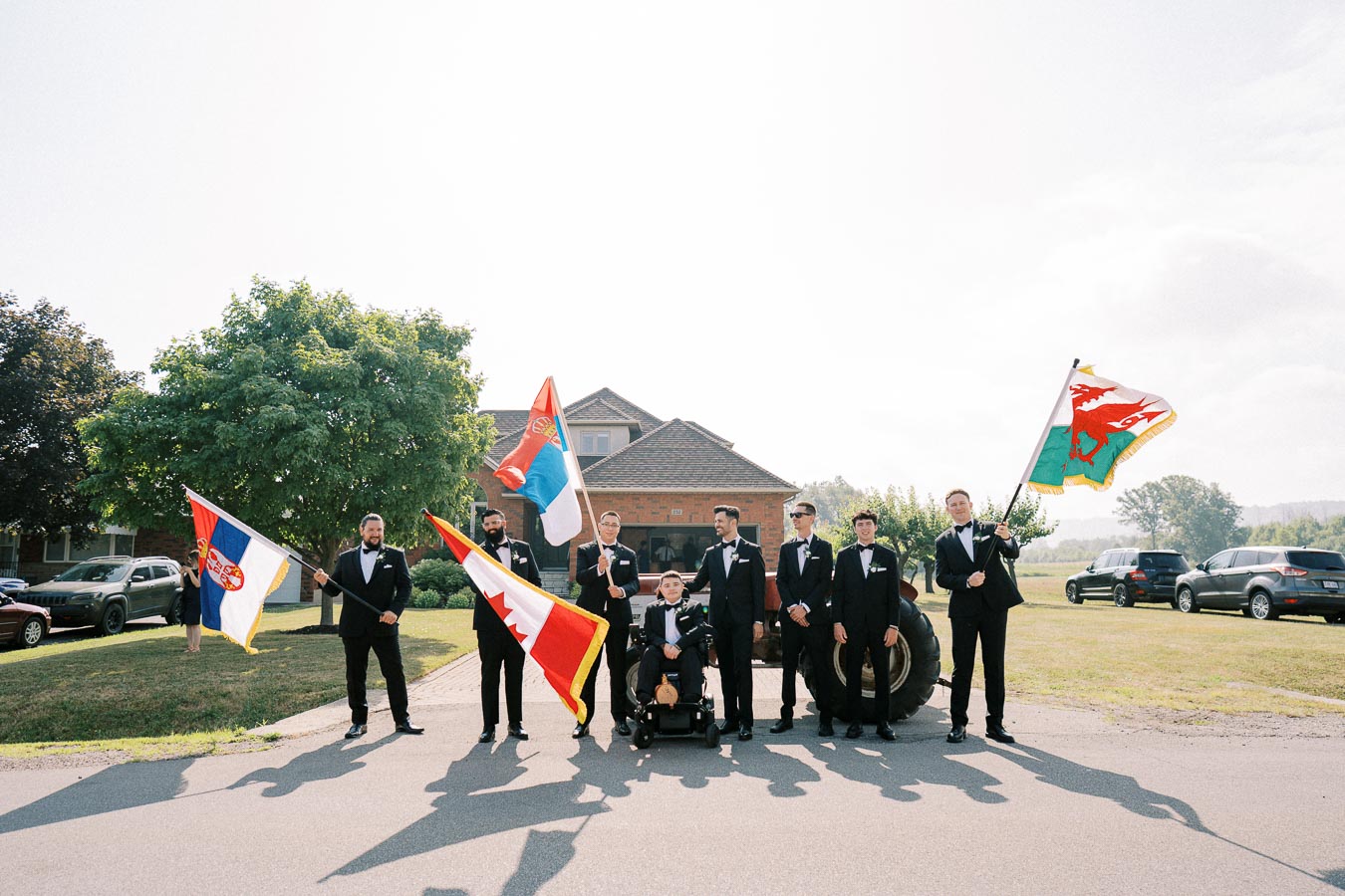 A group of men in formal attire, including tuxedos, stand outside a house holding various national flags on a sunny day, with a backdrop of trees and parked cars.