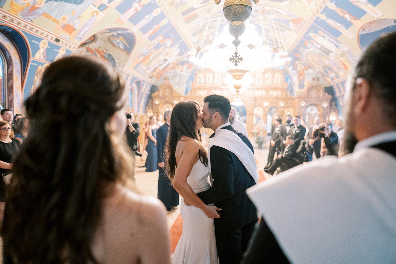Couple sharing a kiss during a traditional wedding ceremony in a beautifully decorated church, surrounded by guests and ornate iconography.