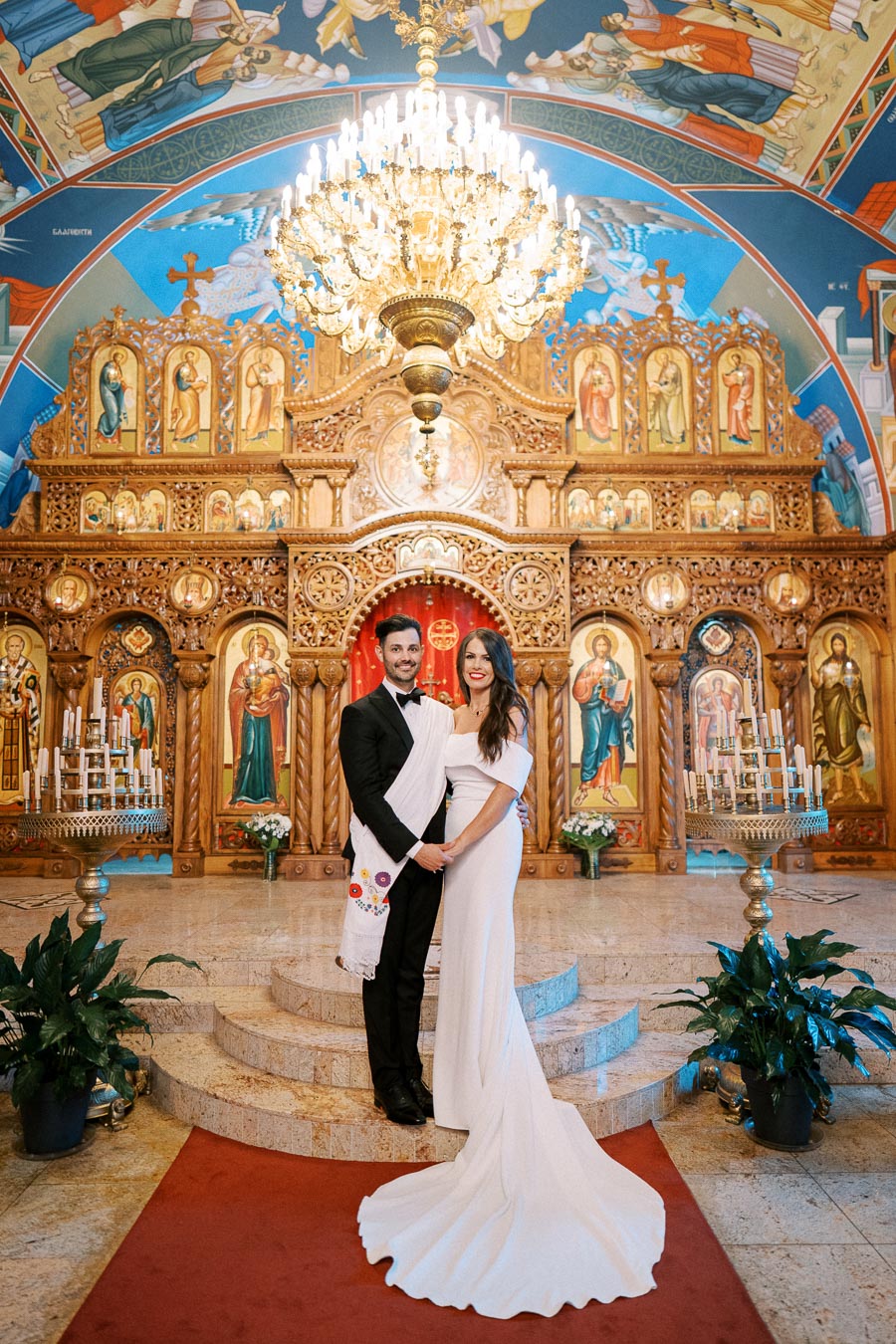 A couple posing in front of an ornate altar inside a beautifully decorated Eastern Orthodox church, adorned with vibrant religious iconography and a grand chandelier, highlighting a traditional wedding ceremony setting.